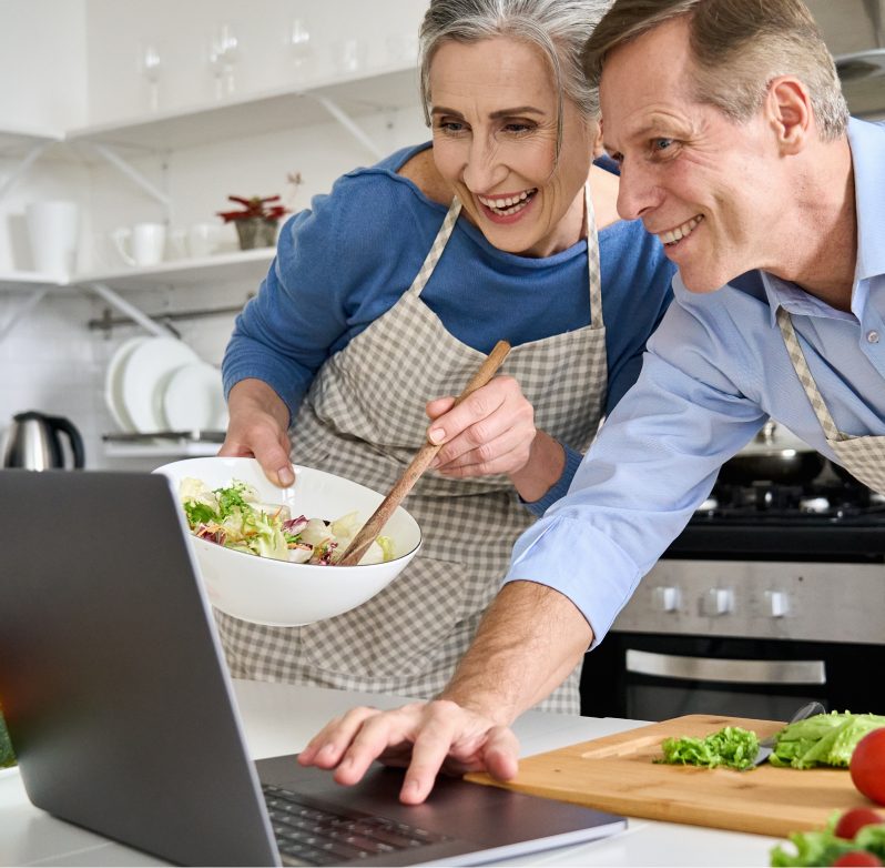 An older couple is cooking together in the kitchen. They are looking at a laptop and laughing.